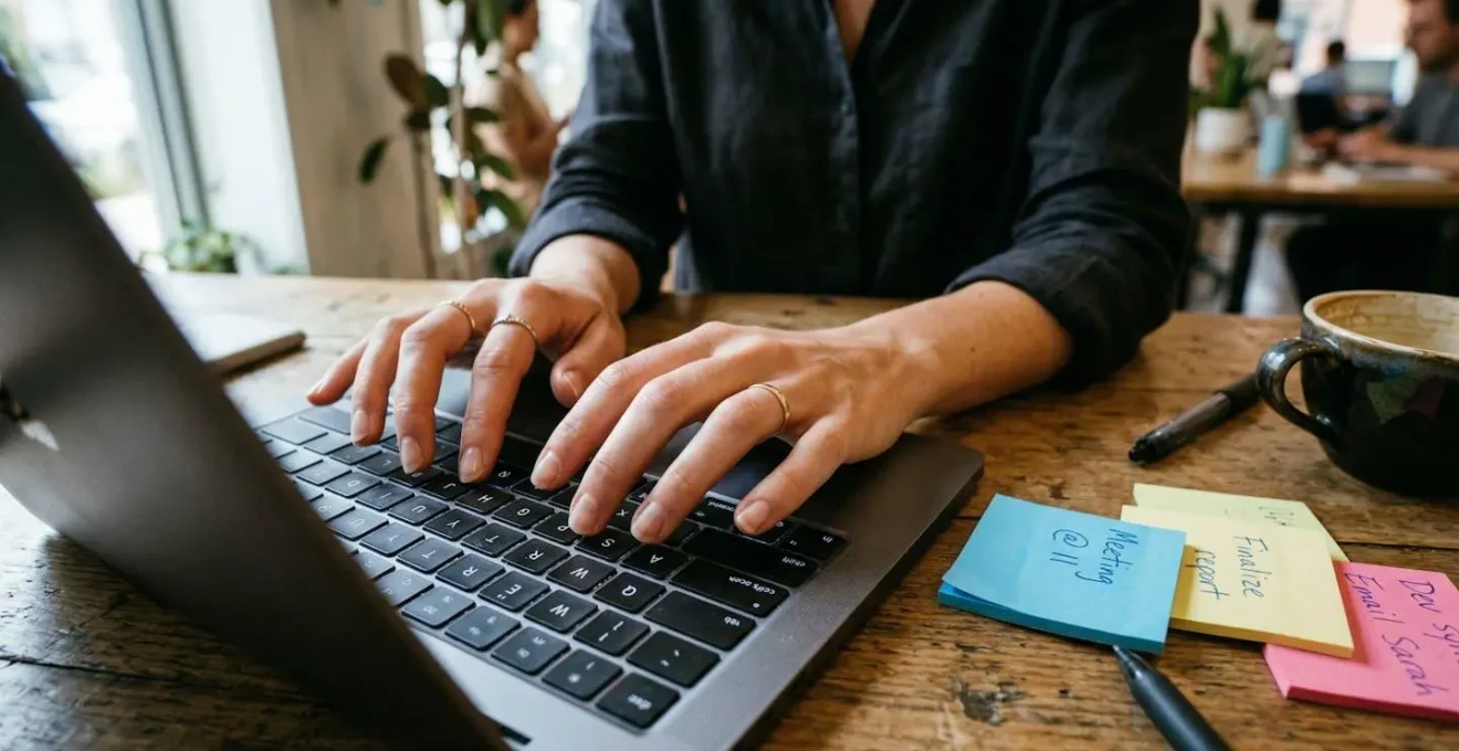 Gros plan sur des mains tapant sur le clavier d'un ordinateur portable moderne, avec des post-it et un carnet visibles en arrière-plan